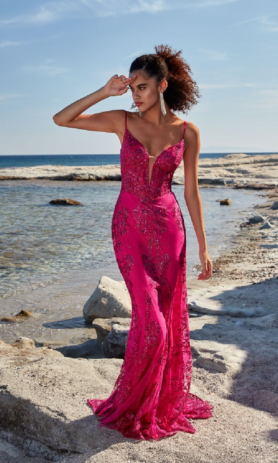 A woman in the Aerie Couture 206 Strappy-Back Long Prom Dress stands on a rocky beach, shielding her eyes from the sun as she gazes into the distance.