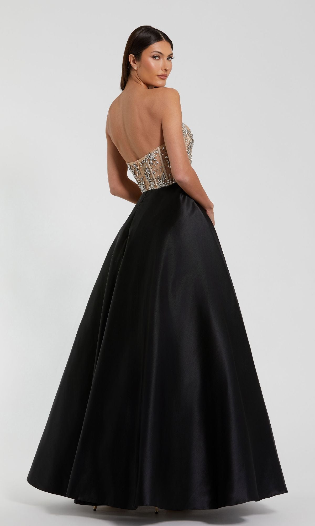 A woman in the Mac Duggal 20701 Strapless Black Prom Ball Gown, featuring a beaded sheer corset-style bodice and full satin skirt, stands facing away and looks over her shoulder.