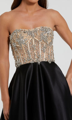 Woman wearing the Strapless Black Prom Ball Gown: Mac Duggal 20701, featuring a nude, beaded and embroidered corset-style bustier bodice with a solid satin A-line skirt, poses with one hand on her waist.
