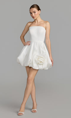 A woman wearing the Mac Duggal 20705 Lilac Bubble-Hem Cocktail Dress in ivory, featuring a strapless silhouette and a large fabric rose detail on the skirt, paired with open-toe heels, stands against a plain gray background—a stunning semi-formal look perfect for special occasions.
