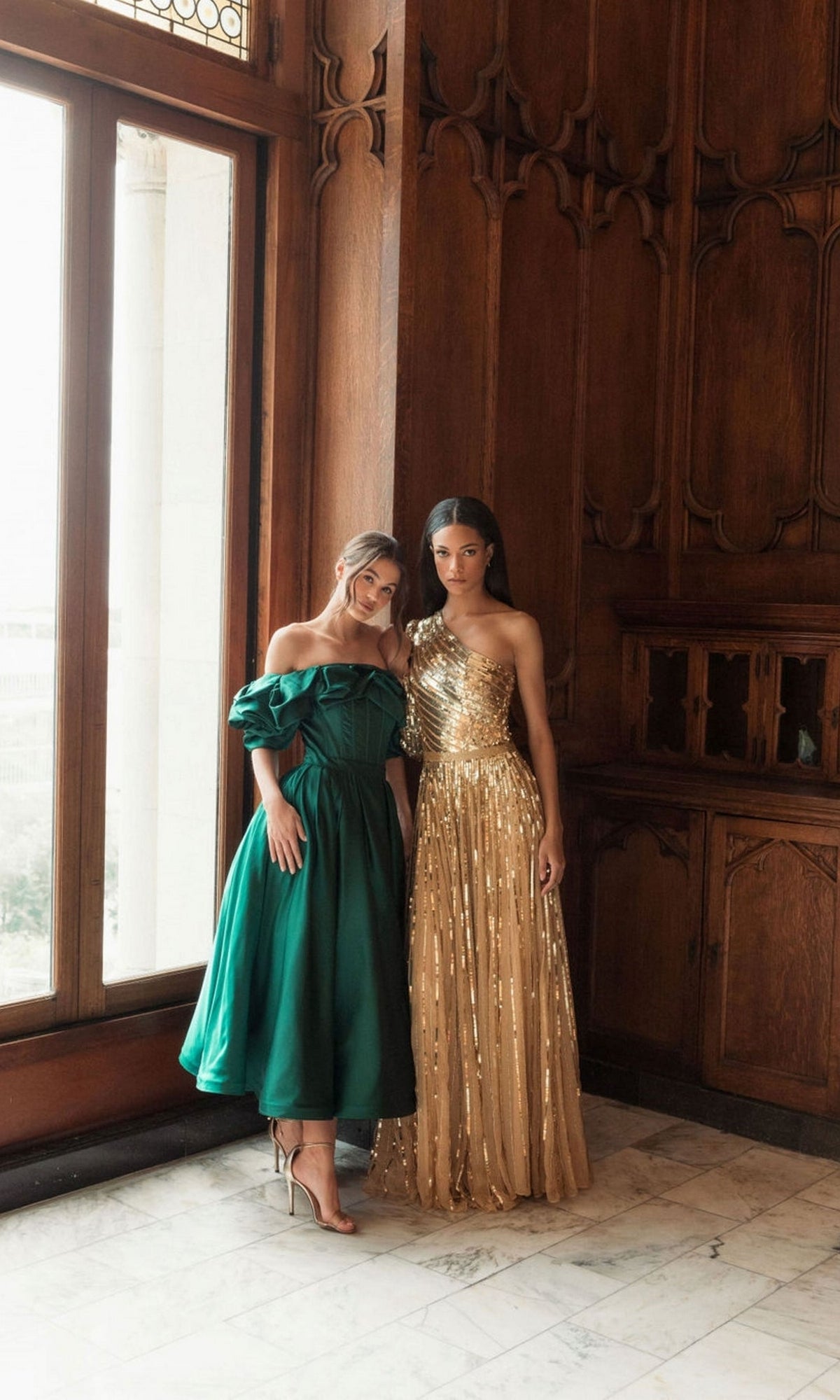 Two women pose indoors against a wood-paneled wall; one wears the green Mac Duggal 20706 off-shoulder wedding guest dress with heels, while the other stuns in a gold one-shoulder floor-length gown.