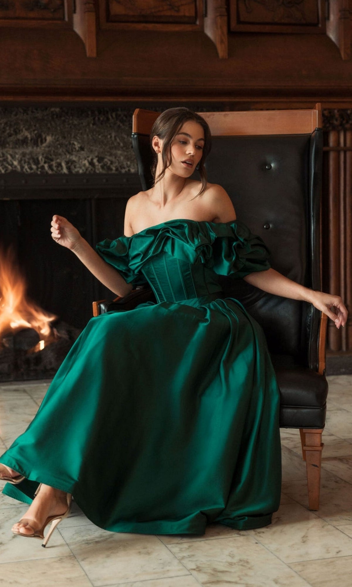 A woman in the Mac Duggal 20706 Off-Shoulder Wedding Guest Dress in emerald green sits on a black leather chair by a fireplace, legs crossed and her gaze turned to the side.