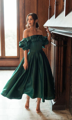 A woman in a green Mac Duggal 20706 off-shoulder wedding guest dress stands by a fireplace in a wood-paneled room with marble floors and large windows.