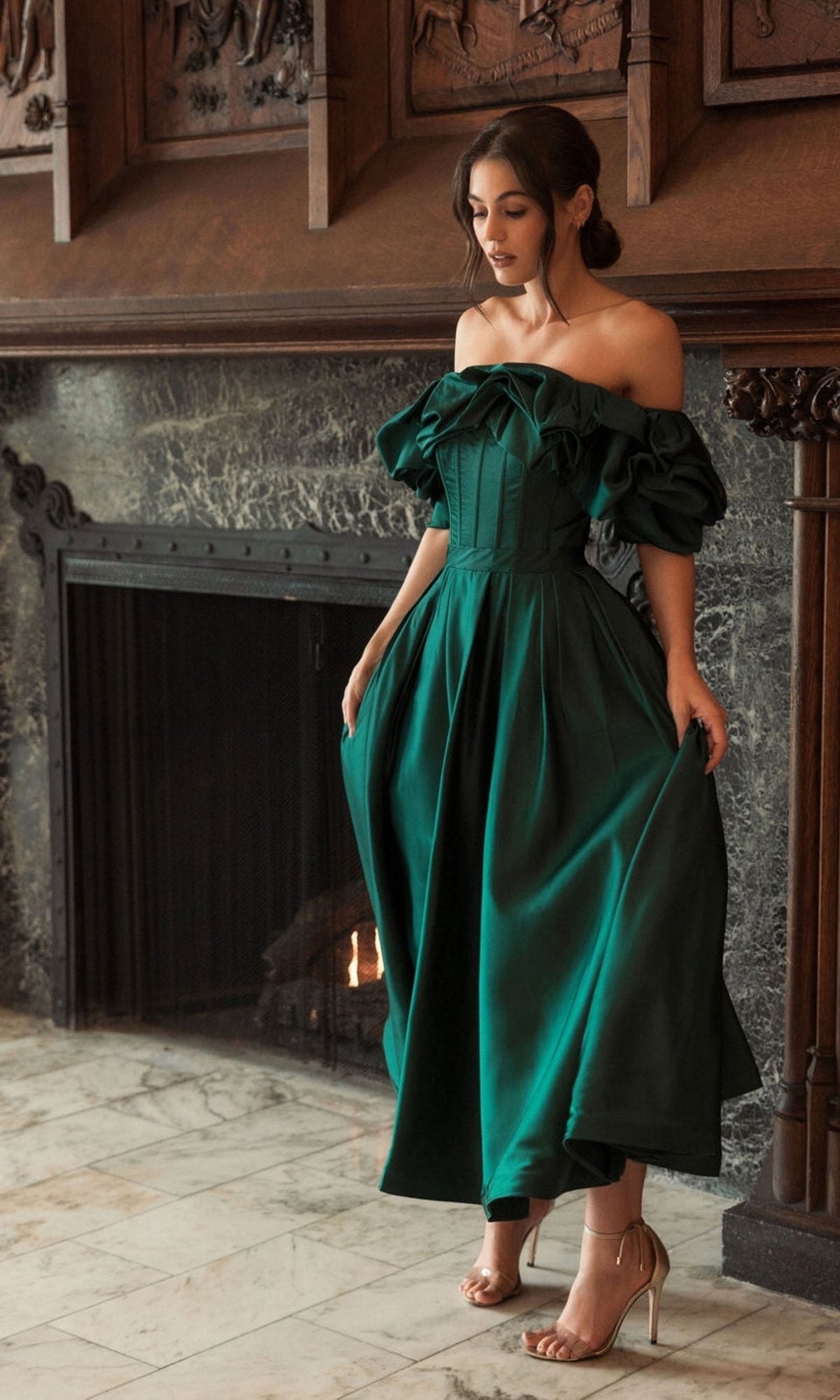 A woman stands by a fireplace, wearing the Off-Shoulder Wedding Guest Dress: Mac Duggal 20706 in emerald green, holding her semi-formal midi dress and looking down, paired with nude heels.