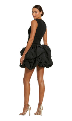A woman models the Short Black Bubble Party Dress (Mac Duggal 20709), featuring a sleeveless design and voluminous mini bubble skirt, standing sideways in high-heeled sandals against a plain white background.