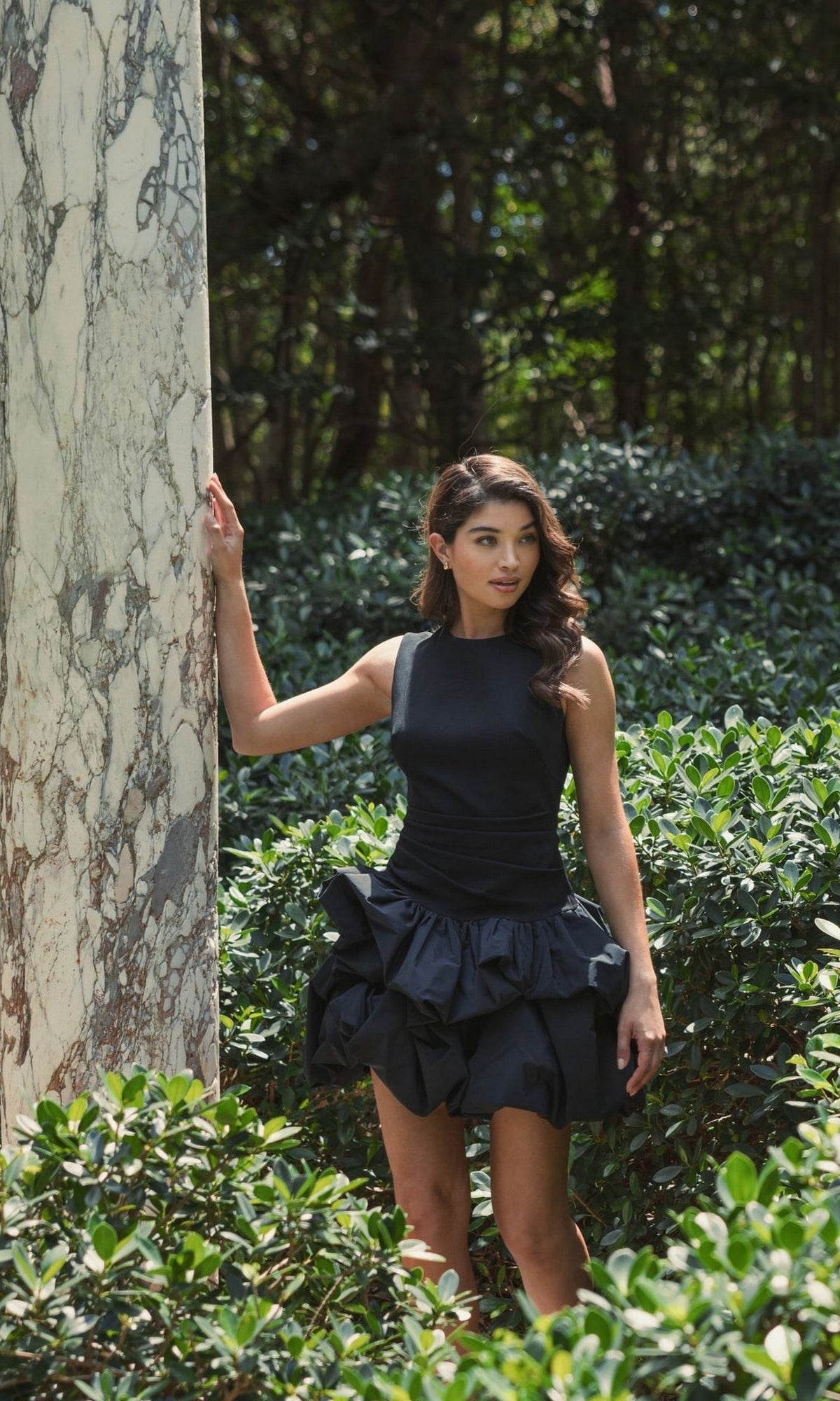 A woman wearing the Mac Duggal 20709 short black bubble party dress poses outdoors by a marble pillar, surrounded by green bushes and trees.