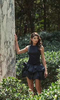 A woman wearing the Mac Duggal 20709 short black bubble party dress poses outdoors by a marble pillar, surrounded by green bushes and trees.