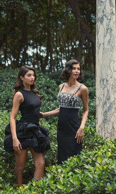 Two women stand in a garden with lush greenery and a marble column; one wears the Short Black Bubble Party Dress, Mac Duggal 20709, while the other dons a sleeveless embellished cocktail dress.