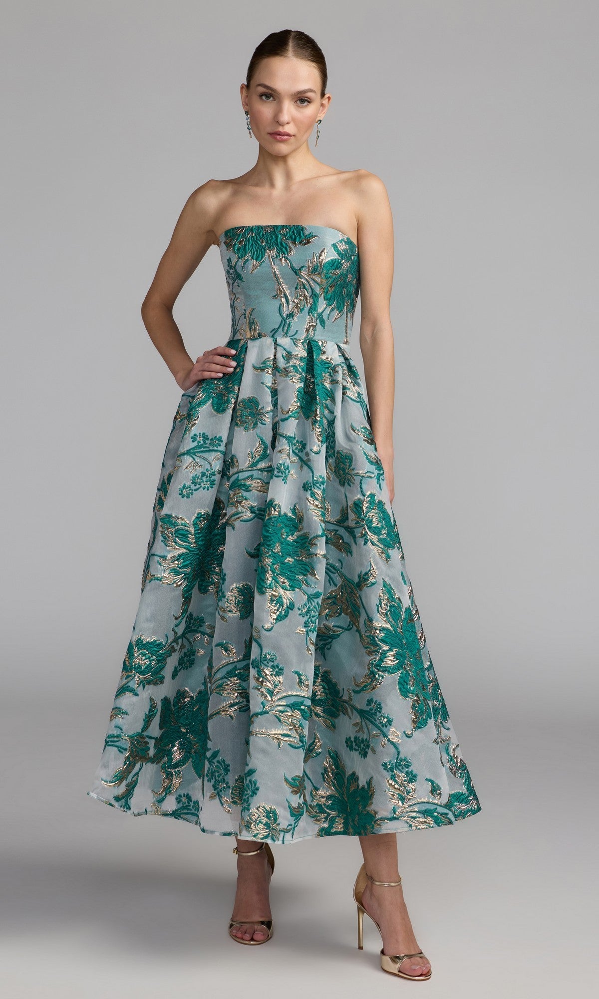 Woman wearing a strapless bronze Mac Duggal 20716 party dress with a flared skirt and gold high heels, standing against a plain gray background.