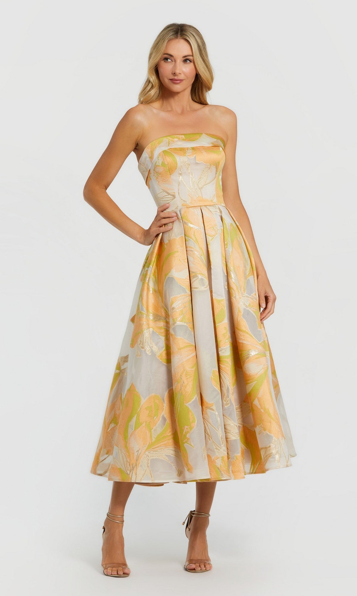 A woman poses with one hand on her hip in the Mac Duggal 20724 Midi Wedding Guest Dress in Peach Multi, featuring a strapless floral design with yellow and orange leaves, styled with nude heels against a plain light background.