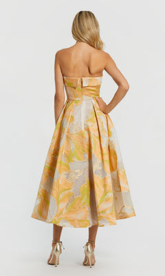 A woman faces away, wearing the strapless Mac Duggal 20724 midi wedding guest dress with a vibrant floral pattern in orange, yellow, and green, paired with metallic high heels.
