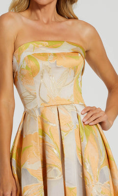 Woman wearing the Mac Duggal 20724 Midi Wedding Guest Dress, a strapless floral design in peach, green, and cream, stands with one hand on her hip.