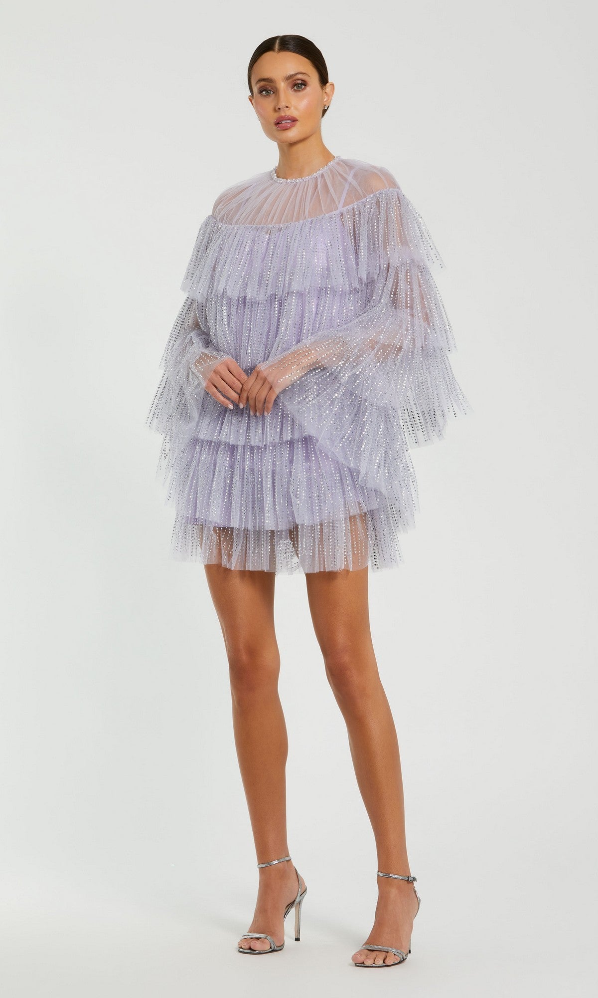 A woman wears the Mac Duggal 20749 short party dress in lilac tulle with a high neck, ruffles, sheer long sleeves, and silver heels, standing against a plain light background.