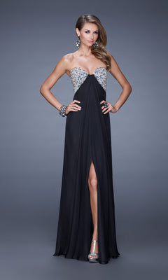 A woman stands wearing the La Femme Dress 20784, a strapless black chiffon gown with an embellished bust and front slit, accessorized with earrings, bracelets, and silver heels.