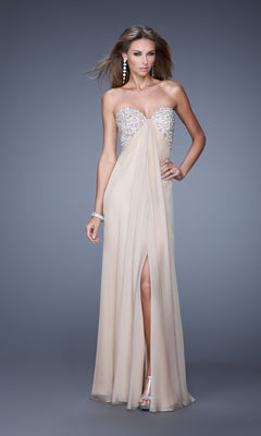 A woman in the La Femme Dress 20784—a strapless, floor-length chiffon gown with an embellished bust and front slit—pairs it with silver high heels, standing against a plain gray background.