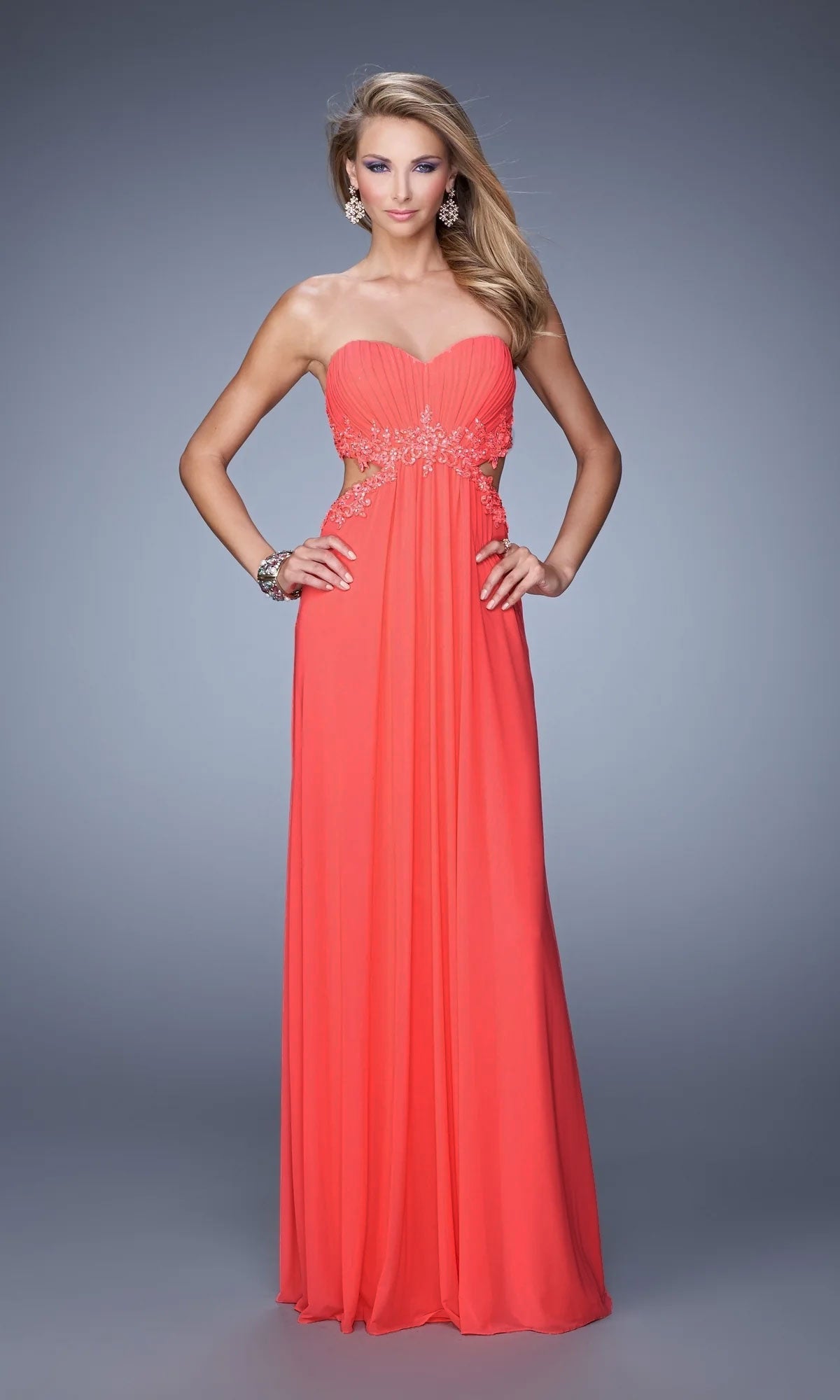 A woman poses against a gray background wearing the Strapless Cut-Out La Femme Jersey Prom Dress 20826 in coral, featuring beaded waist embellishments and delicate beaded lace accents.