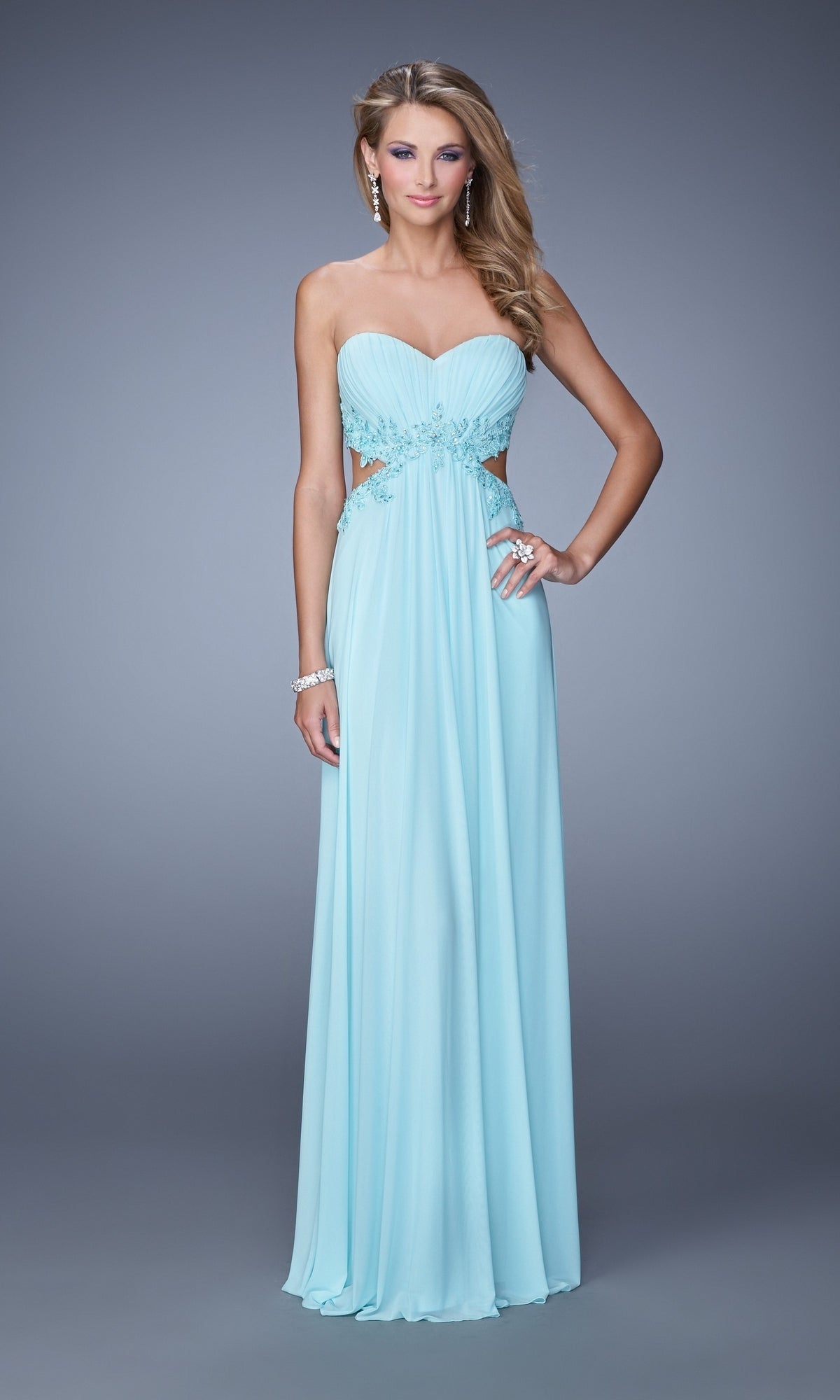 A woman wears the Strapless Cut-Out La Femme Jersey Prom Dress 20826, a light blue, floor-length gown with side cutouts and beaded lace accents, styled with earrings and bracelets against a plain background.