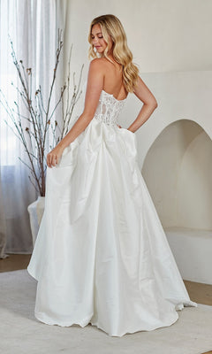 A woman in the Amelia Couture 2087 long prom dress with a strapless lace corset bodice and full A-line skirt stands indoors, glancing over her shoulder.