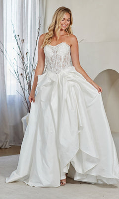 A woman stands indoors, smiling as she holds out the sides of her white Amelia Couture 2087 long prom dress, featuring a strapless lace corset bodice and a full, draped A-line skirt.
