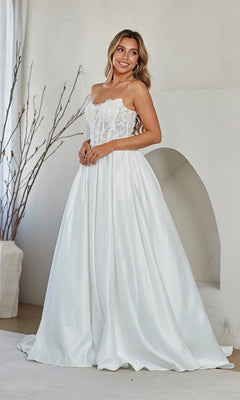 A woman wearing the Amelia Couture 2089 long prom dress with a strapless white lace corset bodice and satin skirt stands indoors near a minimalist archway and decorative branches.
