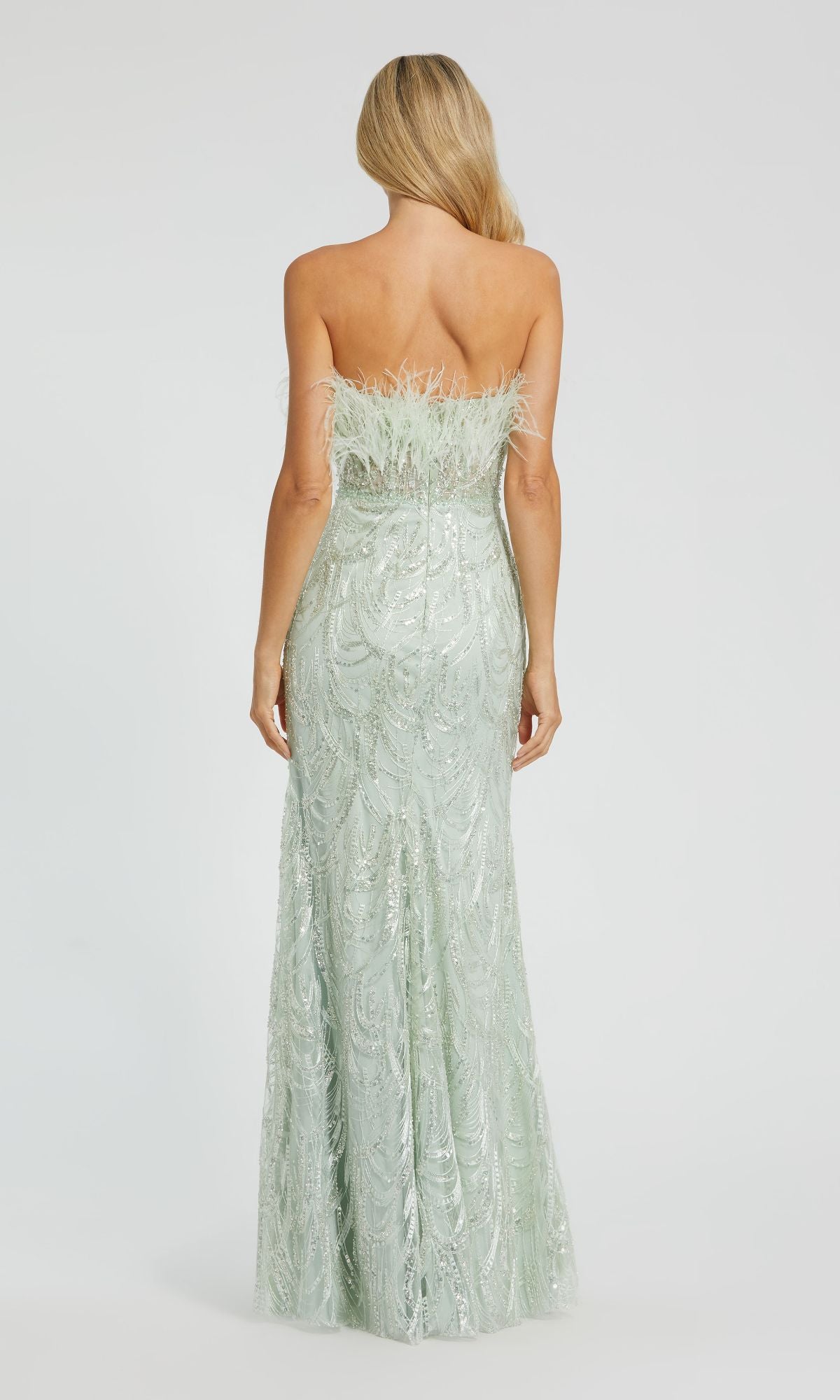 A woman with long blonde hair models the Mac Duggal 20890 long mint sequin prom dress, strapless with feather and sequin details, shown from the back against a plain background.