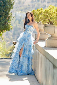 A woman wearing the Long Prom Dress: Amelia Couture 2092, a strapless blue floral dress with a corset bodice and a layered, ruffled skirt with a high leg slit, leaning against a stone wall.