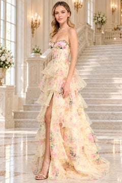 A woman on a marble staircase wearing the Long Prom Dress: Amelia Couture 2092, a strapless, floor-length yellow floral gown with a corset bodice, tiered ruffled skirt, and high side slit.