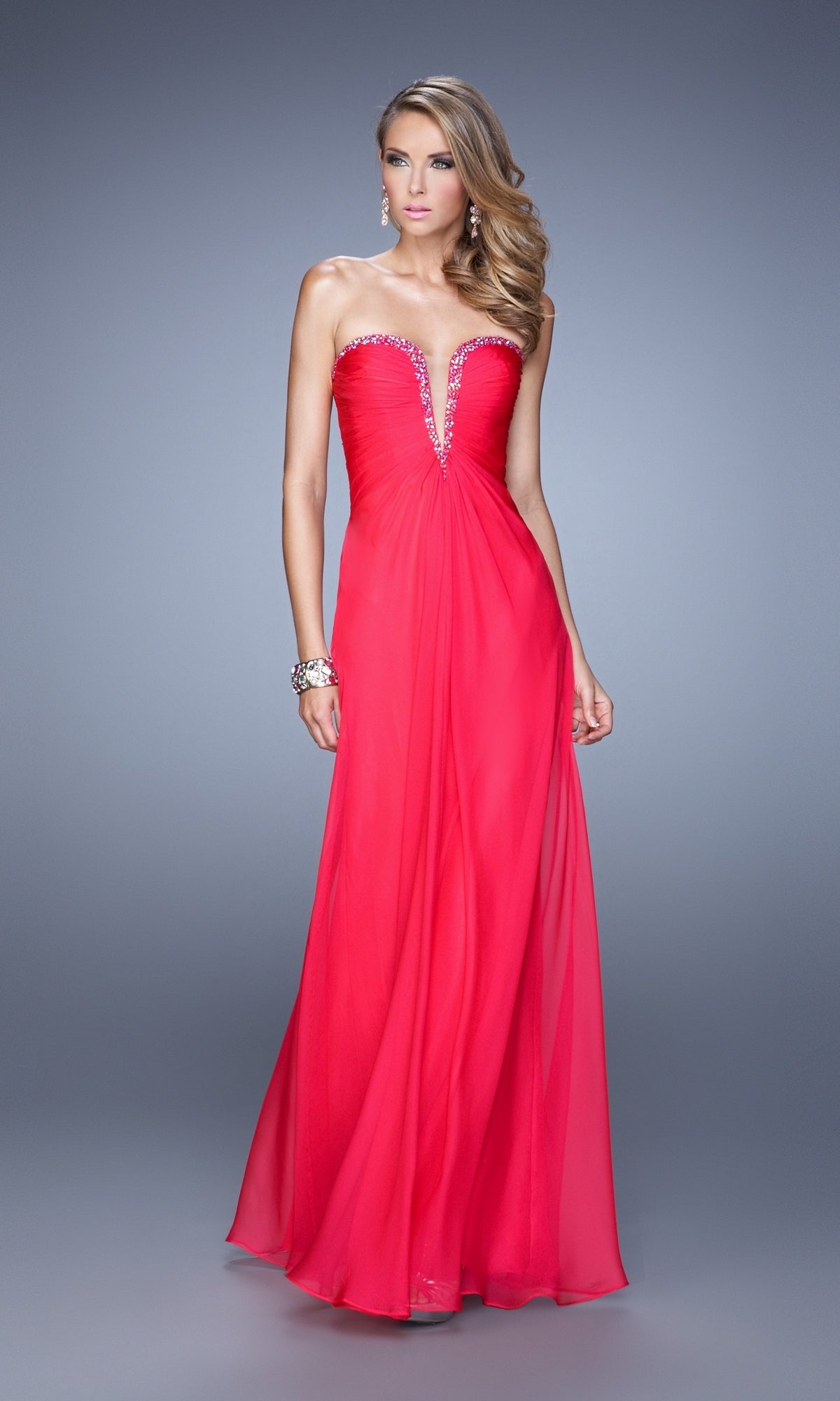 A woman stands against a gray background wearing the La Femme Plunging-Sweetheart Prom Dress 20930, a strapless, floor-length bright pink chiffon gown featuring a crystal-embellished deep V neckline.