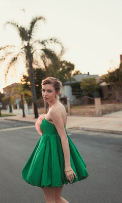 A woman in a Mac Duggal 20931 Short Bubble Party Dress—strapless, green, with a pink flower accent—stands on a residential street at sunset, glancing back over her shoulder.