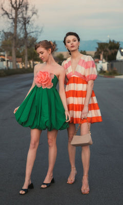 Two women stand on a street; one wears the Mac Duggal 20931 Short Bubble Party Dress with a large pink flower, while the other sports a pink and orange striped semi-formal dress with puff sleeves, carrying a beige handbag.