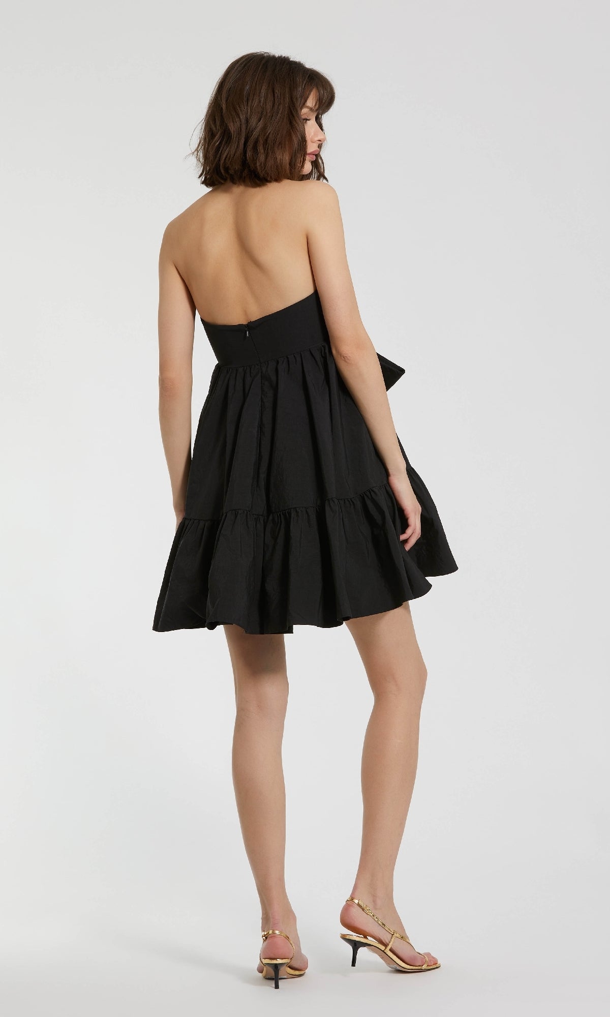 A woman faces away, wearing the Strapless Short Bow Party Dress: Mac Duggal 20937 with a tiered skirt and light heeled sandals, standing against a plain white background.