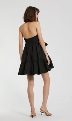 A woman faces away, wearing the Strapless Short Bow Party Dress: Mac Duggal 20937 with a tiered skirt and light heeled sandals, standing against a plain white background.