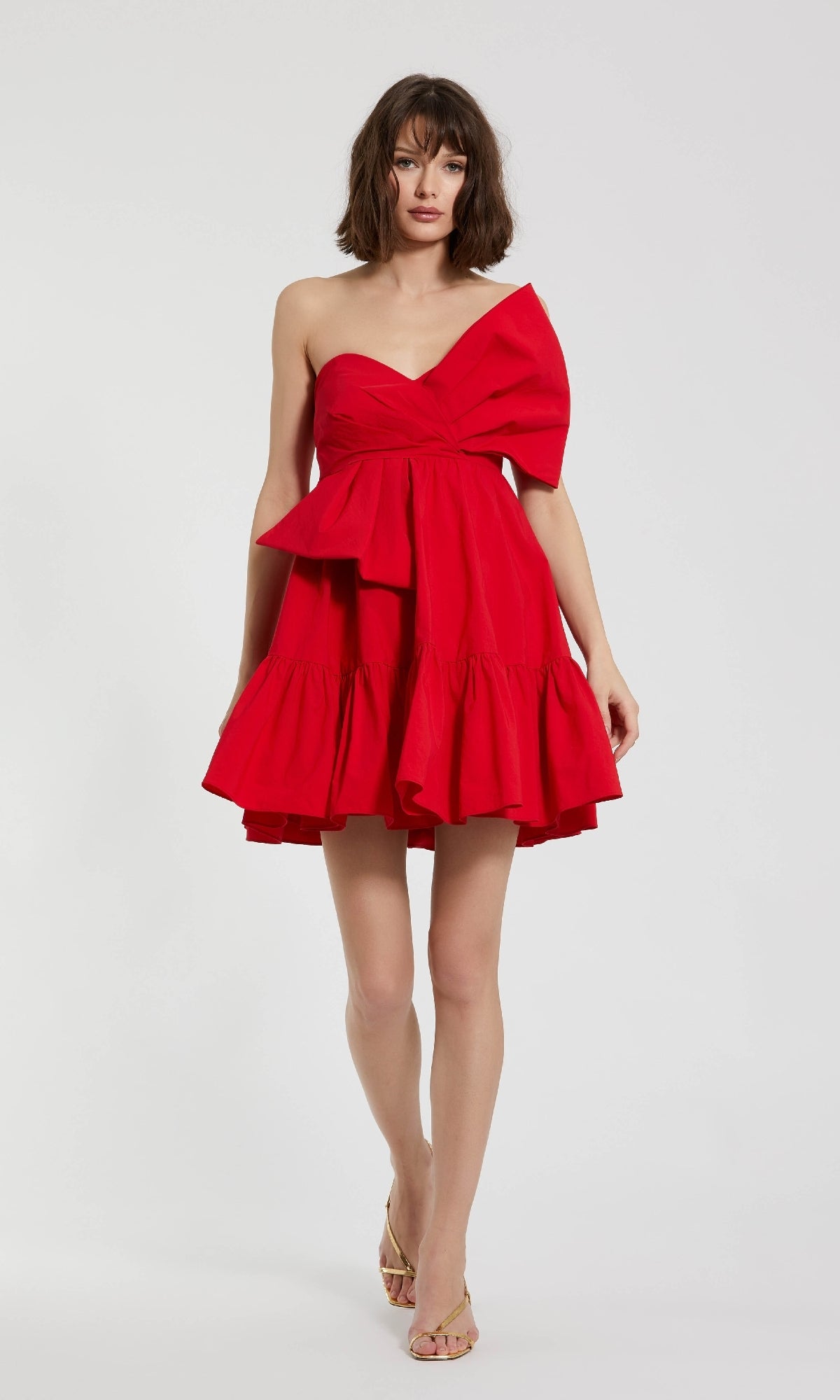 A woman stands against a plain white background wearing the Mac Duggal 20937 Strapless Short Bow Party Dress in bright red with a large bow and ruffled hem.