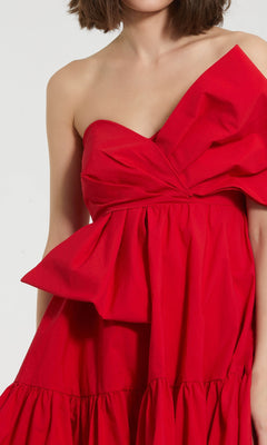 A woman wears the Mac Duggal 20937 Strapless Short Bow Party Dress in red, featuring a large bow detail and a ruffled skirt.