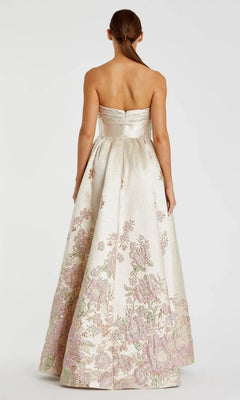 A woman with straight brown hair in a ponytail stands elegantly with her back to the camera, wearing the Mac Duggal 20974—a strapless, floor-length cream floral ballgown with pink and green patterns.