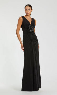 A woman stands against a plain light background wearing the Mac Duggal 21005, a sleeveless, floor-length black formal dress featuring a V-neckline, ruched detailing, and a 3D flower accent at the waist.