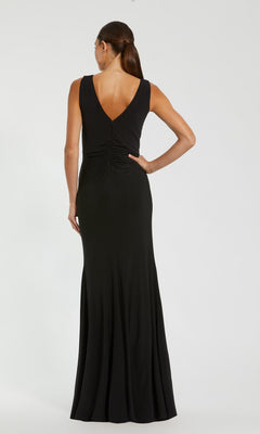 A woman wearing the Mac Duggal 21005 long formal dress—a sleeveless, floor-length black ruched gown with a deep V-back—stands against a plain light background, facing away from the camera.
