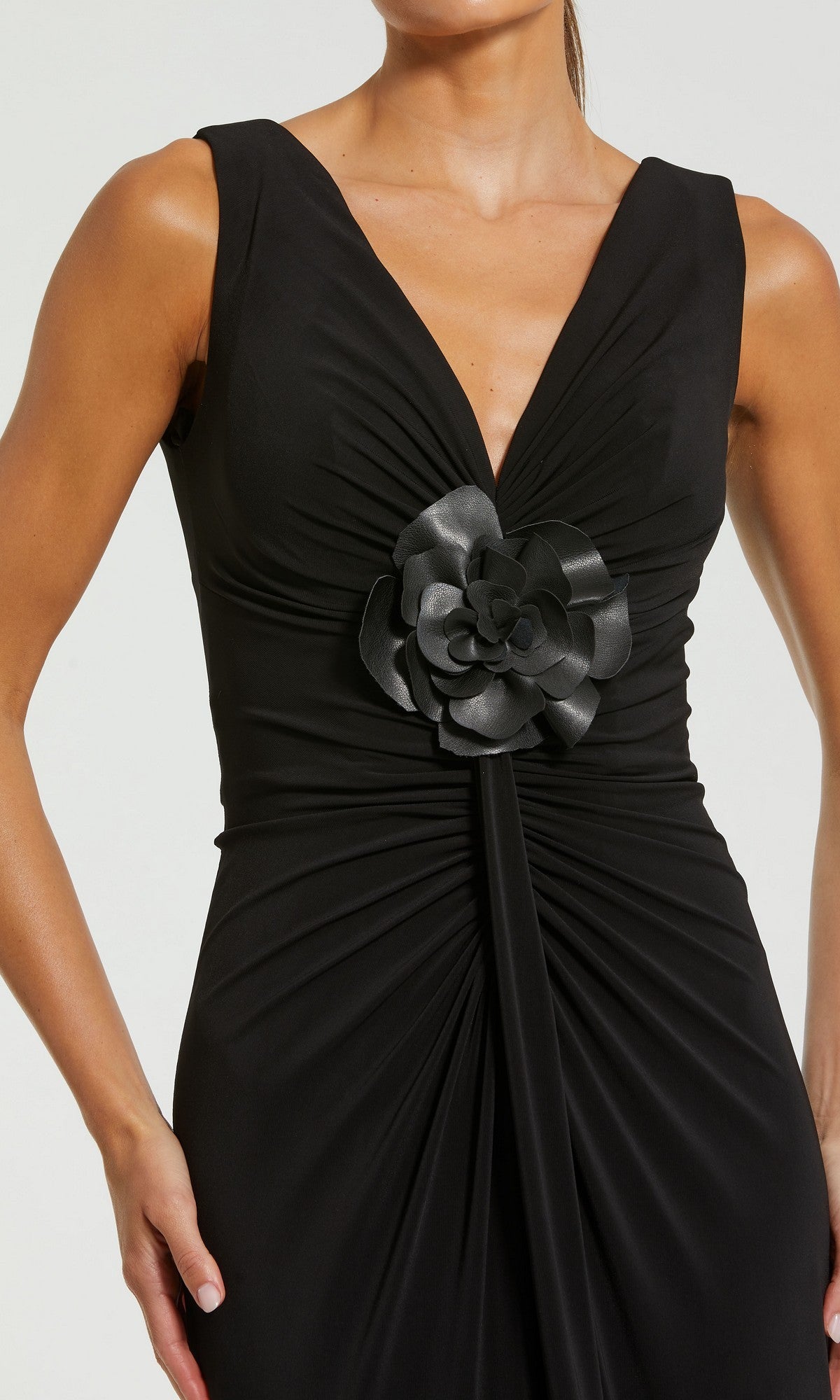 A woman wearing the Mac Duggal 21005 long formal dress, featuring a sleeveless black design with a deep V-neckline, ruched detailing, and a dramatic 3D flower accent at the waist.
