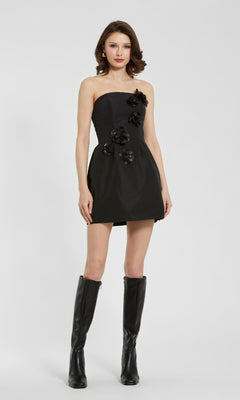 A woman stands against a plain background wearing the Mac Duggal 21008 Strapless Black Cocktail Dress with flower appliqués and knee-high black boots.