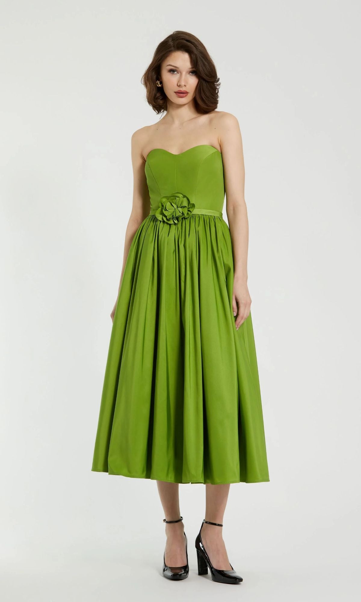 Woman in the Mac Duggal 21023 midi strapless semi-formal dress, green with a fitted bodice, flared tea-length skirt, and waist floral detail—styled with black heels—a chic pick for any semi-formal event.
