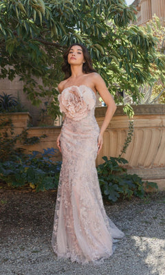 A woman stands outdoors in the Mac Duggal 21036 Strapless Long Floral Prom Dress, featuring a pale mocha pink hue, a large floral embellishment on the bodice, and delicate lace details for an elegant evening look.