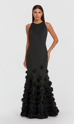 A woman wears the Mac Duggal 21038 long formal dress—sleeveless, floor-length, black with 3D floral appliqués on the lower half and a subtle mermaid silhouette—standing against a plain light background.