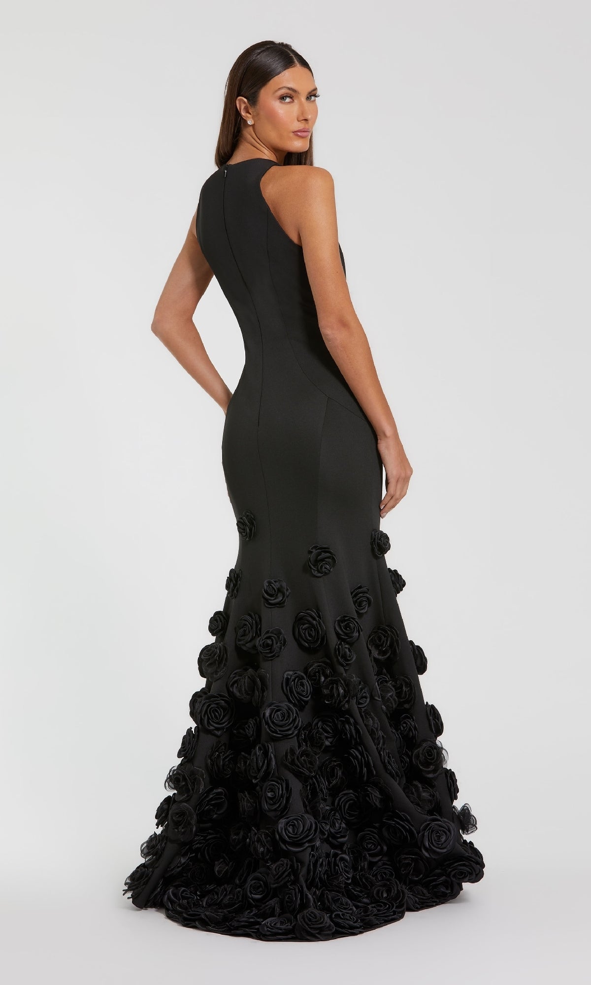 A woman models the Mac Duggal 21038, a sleeveless black floor-length evening gown with 3D floral appliqués on the skirt and a mermaid silhouette, posing against a plain, light background and gazing over her shoulder.