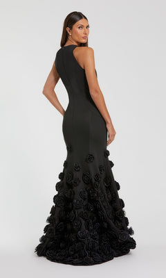A woman models the Mac Duggal 21038, a sleeveless black floor-length evening gown with 3D floral appliqués on the skirt and a mermaid silhouette, posing against a plain, light background and gazing over her shoulder.