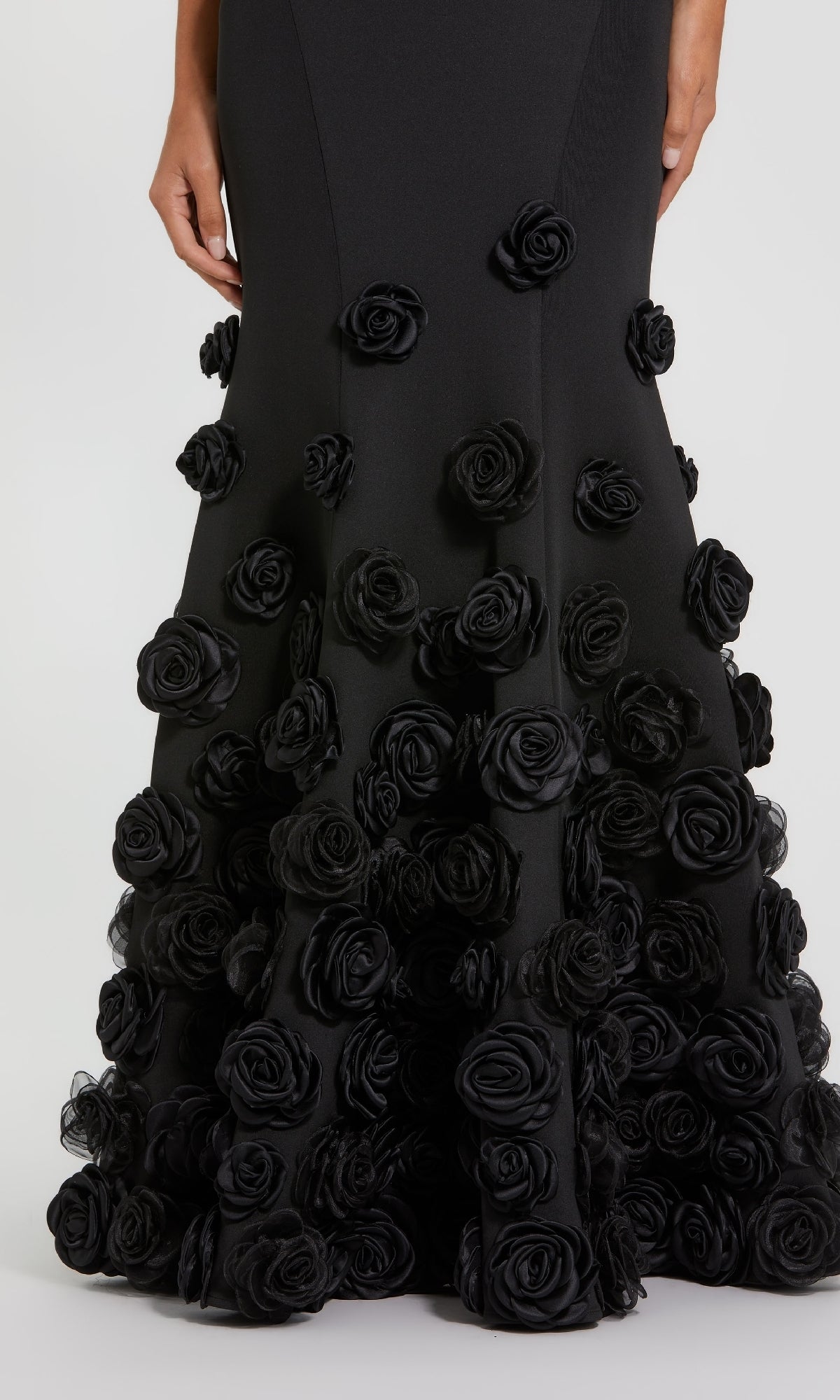 A person is wearing the Mac Duggal 21038 long formal dress, a designer evening gown with a mermaid silhouette and three-dimensional black fabric roses embellishing the lower hem.