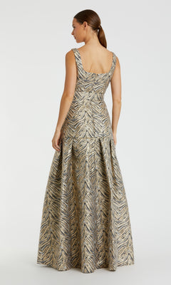 A woman stands facing away, wearing the Mac Duggal 21054 Gold Zebra-Print Long Prom Gown featuring a metallic pattern and pleated skirt.