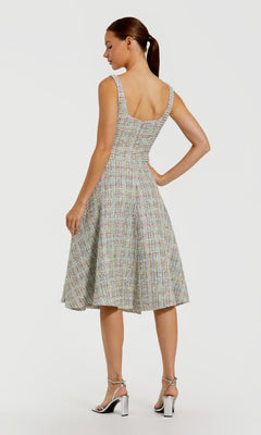 A woman stands facing away in the Mac Duggal 21066 Midi Party Dress, a sleeveless boucle design with a square neckline, paired with silver high-heeled sandals, set against a plain light background.