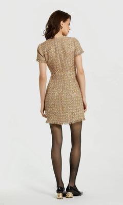 A woman faces away, wearing the Short Wedding Guest Dress: Mac Duggal 21072— a boucle mini sheath with short sleeves—paired with black tights and black shoes featuring gold accents, against a plain light background.