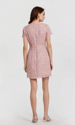 A woman faces away, wearing the Mac Duggal 21072 Short Wedding Guest Dress in pink with short sleeves and white sandals, set against a plain light background.