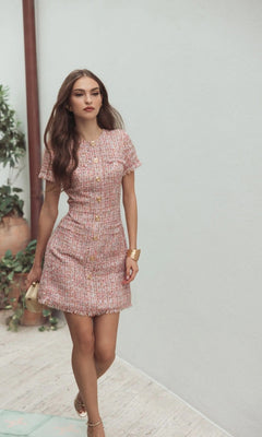 A woman with long brown hair wears the pink Mac Duggal 21072 short wedding guest dress with a high neckline and gold buttons, paired with beige heels, walking indoors by a white wall and green plants.